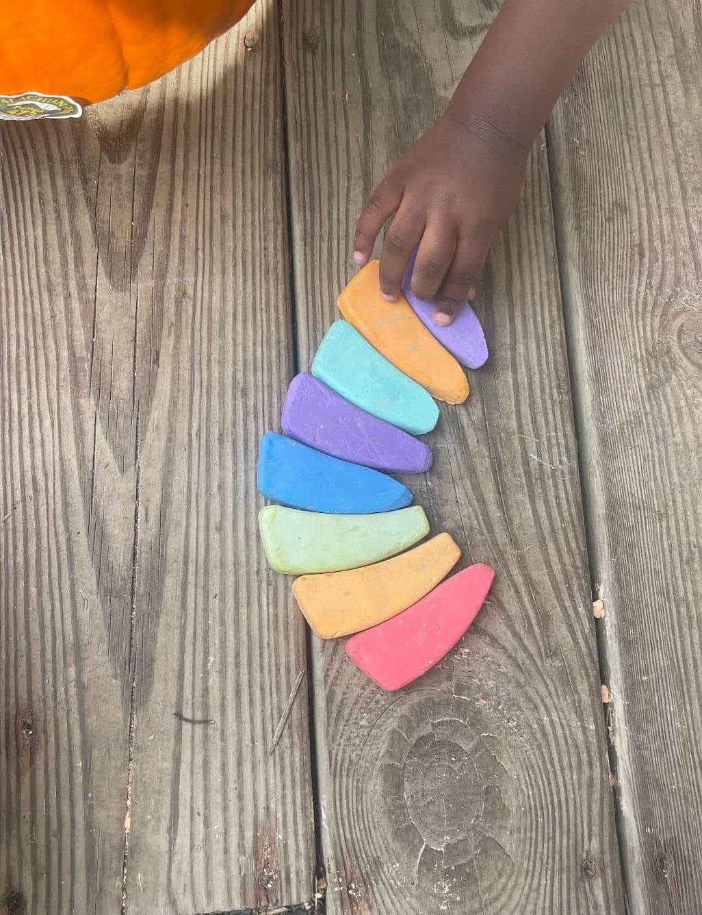 A child's hand lining up rainbow sidewalk chalk on a wooden deck